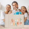A beige tote bag with the text "GROOVY MAMA" printed on it, along with various colorful flowers and peace signs, is displayed in the foreground. In the background, there are two smiling women, one of whom is holding a bouquet of flowers.