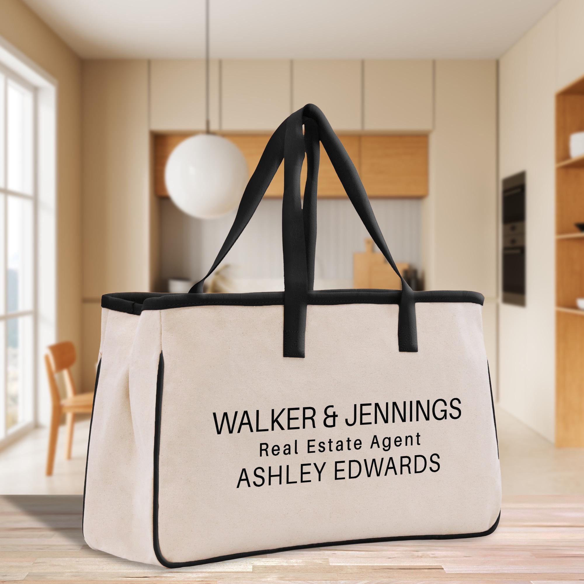 A large canvas tote bag with the text &quot;WALKER & JENNINGS Real Estate Agent ASHLEY EDWARDS&quot; printed on it, placed on a wooden floor in a room with a window and a wooden chair.