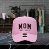 A pink baseball cap with the text "MOM EST. 2025" printed on it, placed on a marble countertop.
