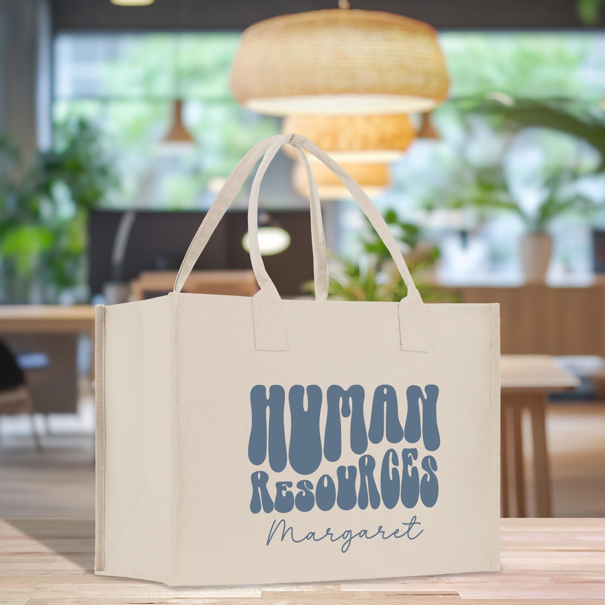 A white tote bag with the text &quot;HUMAN RESOURCES Margaret&quot; printed on it, placed on a wooden surface in a room with a window and plants in the background.