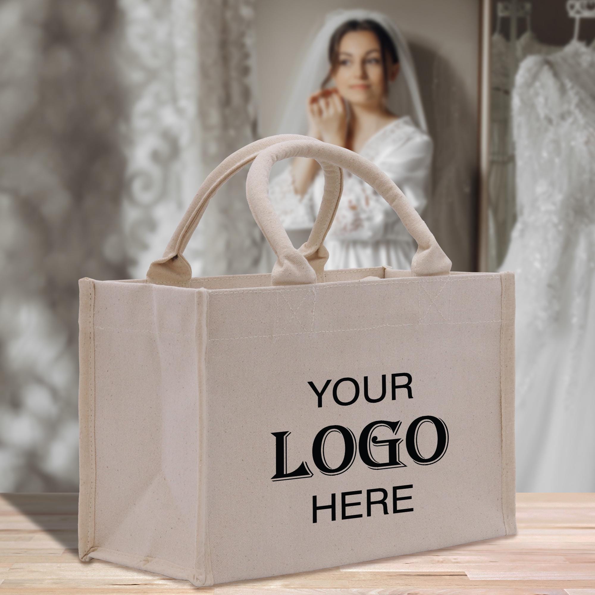 A beige tote bag with the text &quot;YOUR LOGO HERE&quot; printed on it, placed on a wooden surface. In the background, there is a blurred image of a woman wearing a white dress, possibly a bride, and a mirror.
