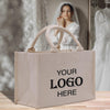 A beige tote bag with the text "YOUR LOGO HERE" printed on it, placed on a wooden surface. In the background, there is a blurred image of a woman wearing a white dress, possibly a bride, and a mirror.