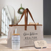a beige tote bag with the name "Olivia Williams" printed on it, along with contact information for a real estate agent, including a phone number and website. Next to the bag is a clear glass jar with a straw, and a small metal container with a lid. The background includes a window and a plant, creating a homely atmosphere.