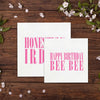 two white greeting cards with pink text, placed on a wooden surface with white flowers scattered around.