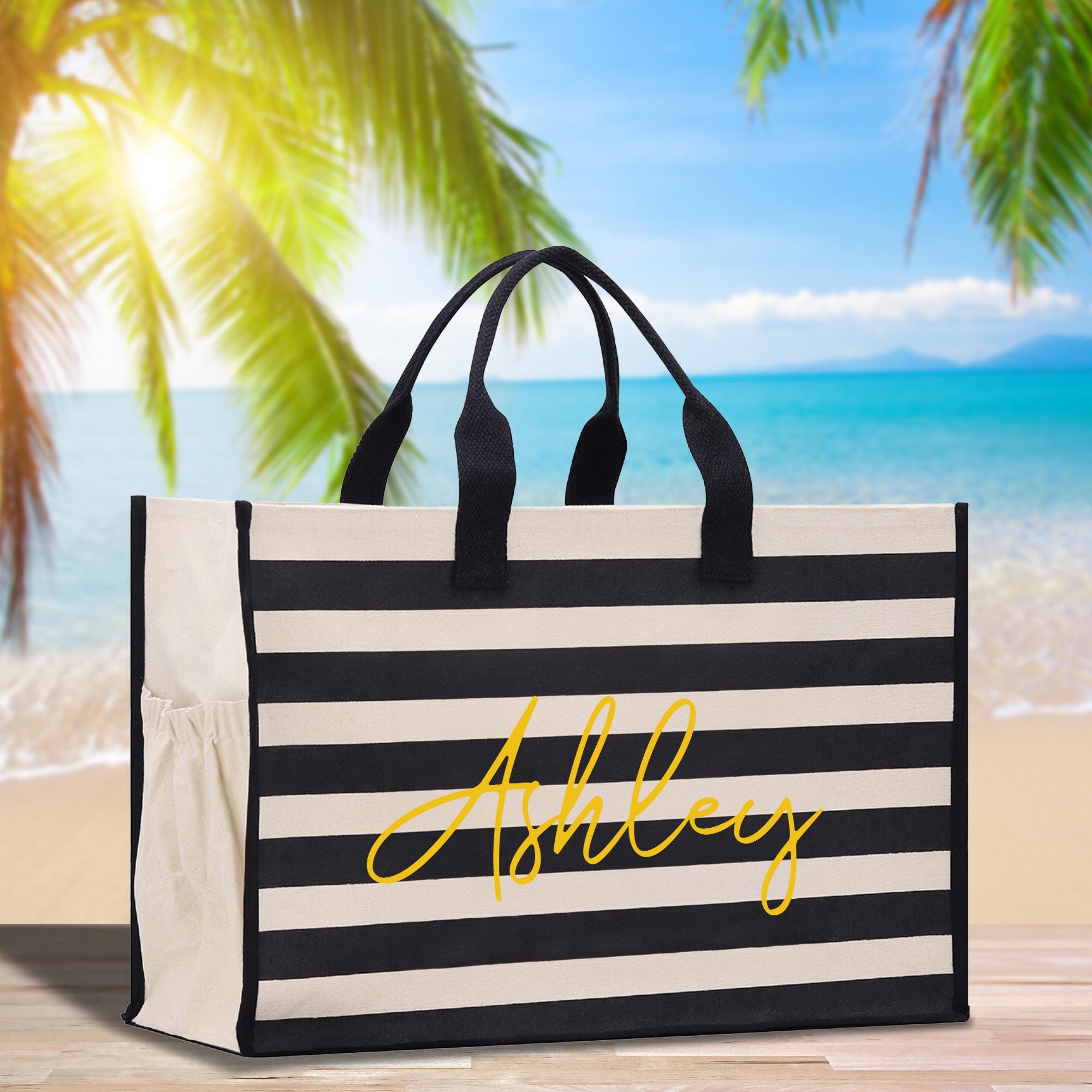 A black and white striped tote bag with the name &quot;Ashley&quot; written in yellow cursive on the front, placed on a wooden surface with palm trees and a beach in the background.
