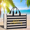 A black and white striped tote bag with the name "Ashley" written in yellow cursive on the front, placed on a wooden surface with palm trees and a beach in the background.