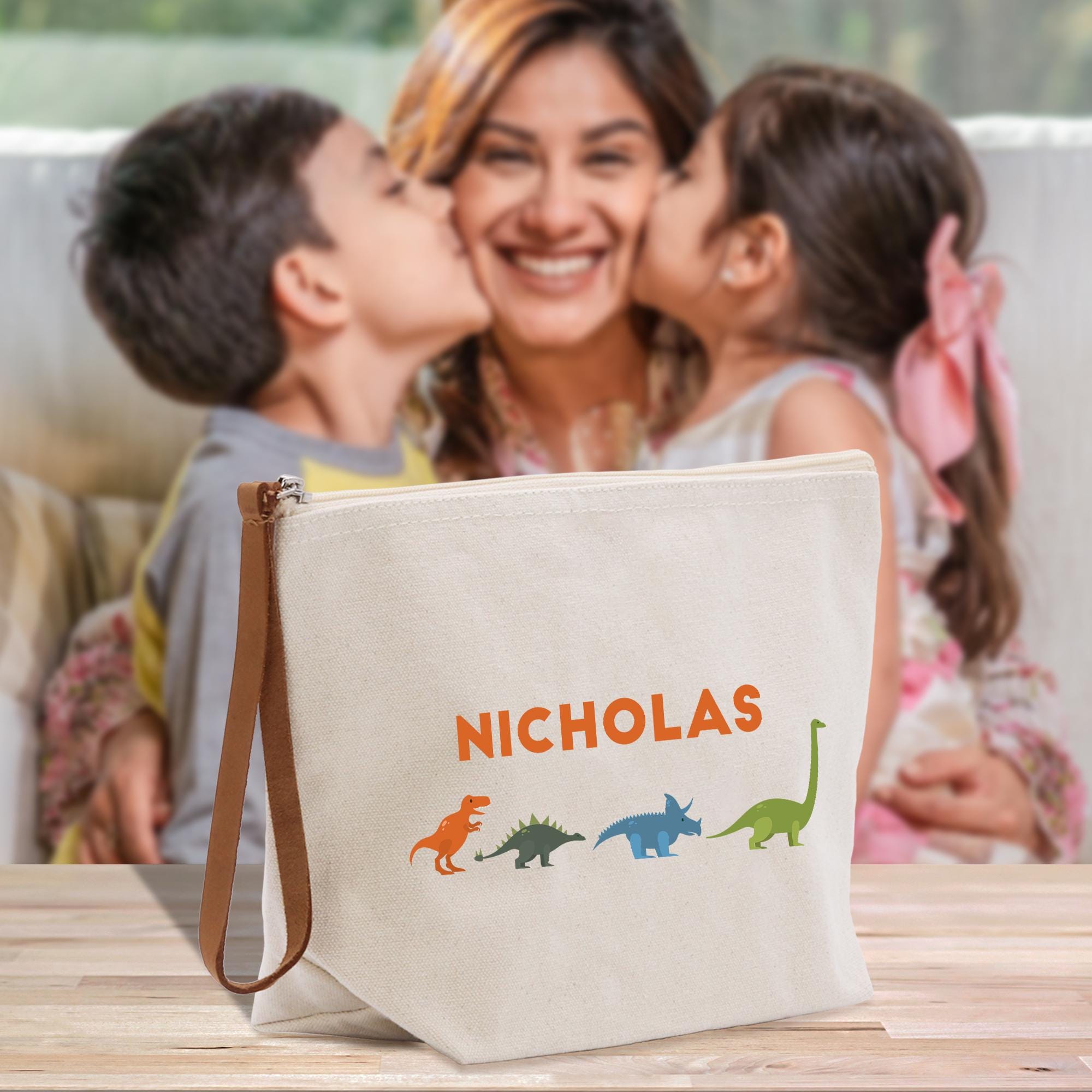 A woman is kissing two children on the cheek, and there is a white bag with a dinosaur design in front of them.