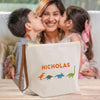 A woman is kissing two children on the cheek, and there is a white bag with a dinosaur design in front of them.