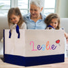 A woman and two young girls are sitting together, looking at a large bag with the name "Leslie" printed on it.