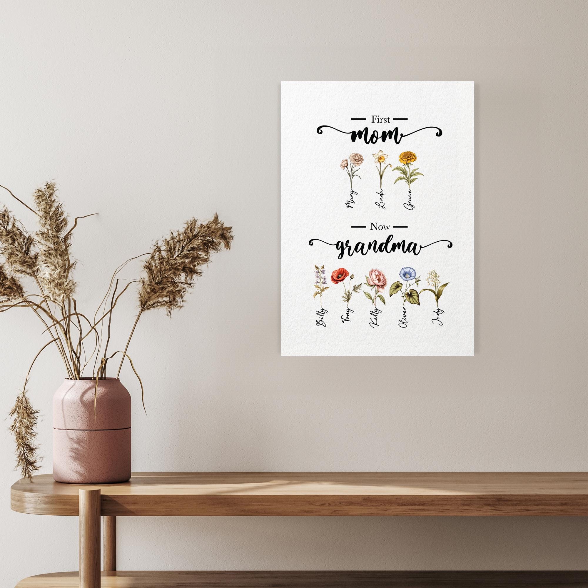 a framed print hanging on a wall, depicting a floral design with the text &quot;First Mom Now Grandma&quot; written above and below the flowers.