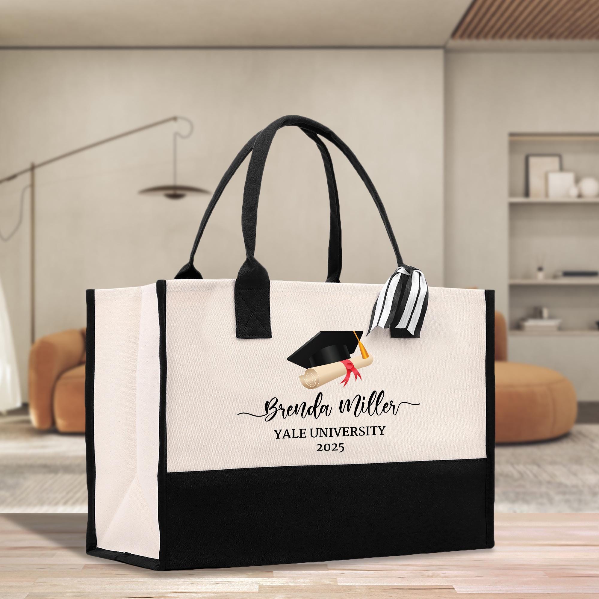 A large tote bag with a graduation cap and mortarboard printed on it, placed on a wooden floor in a room with a brown sofa and a bookshelf in the background.