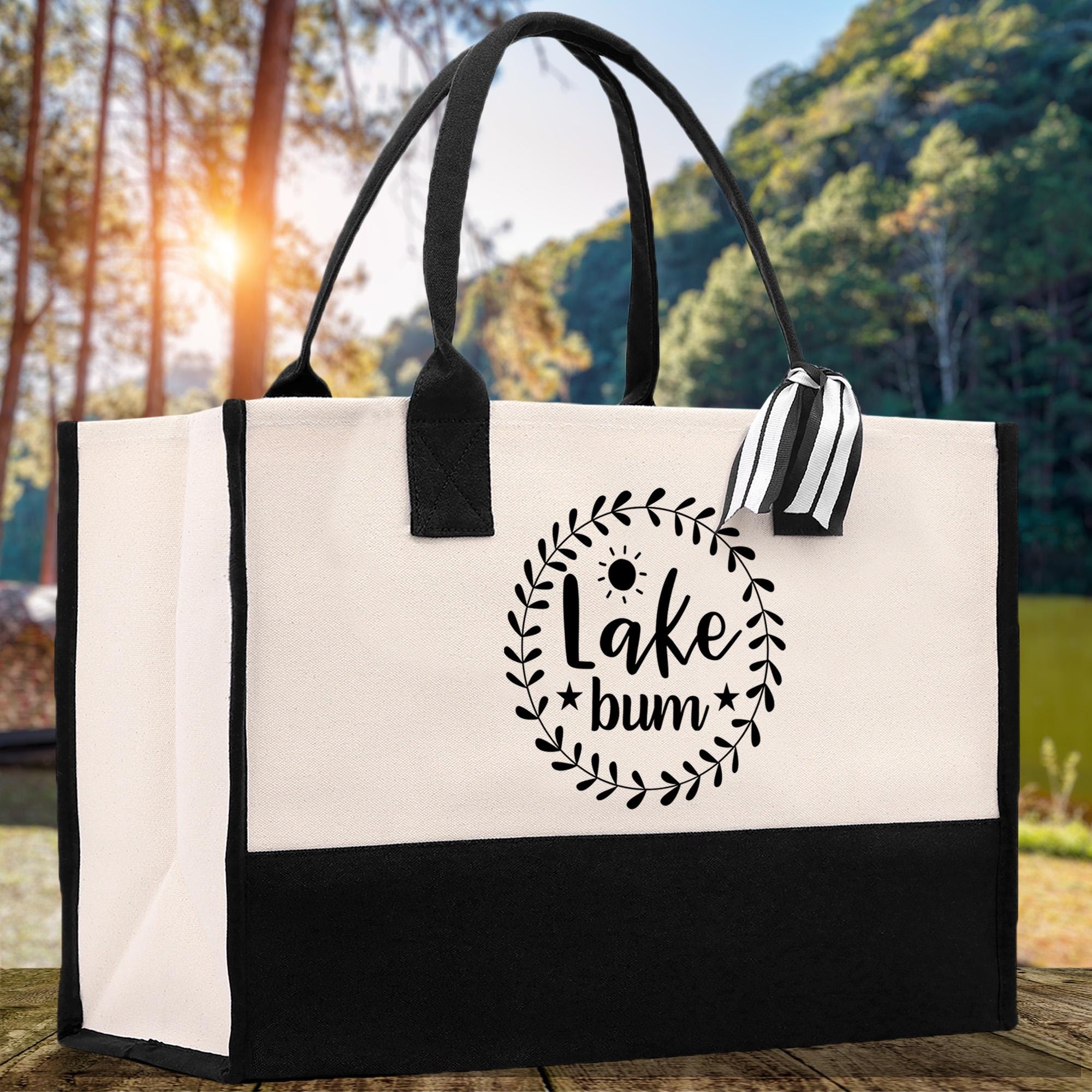 A black and white tote bag with the text "Lake + Bum" printed on it, placed on a wooden surface with a natural outdoor background.