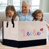 A woman and two young girls are sitting together on a couch, with a large tote bag with the name "Leslie" printed on it in the foreground.