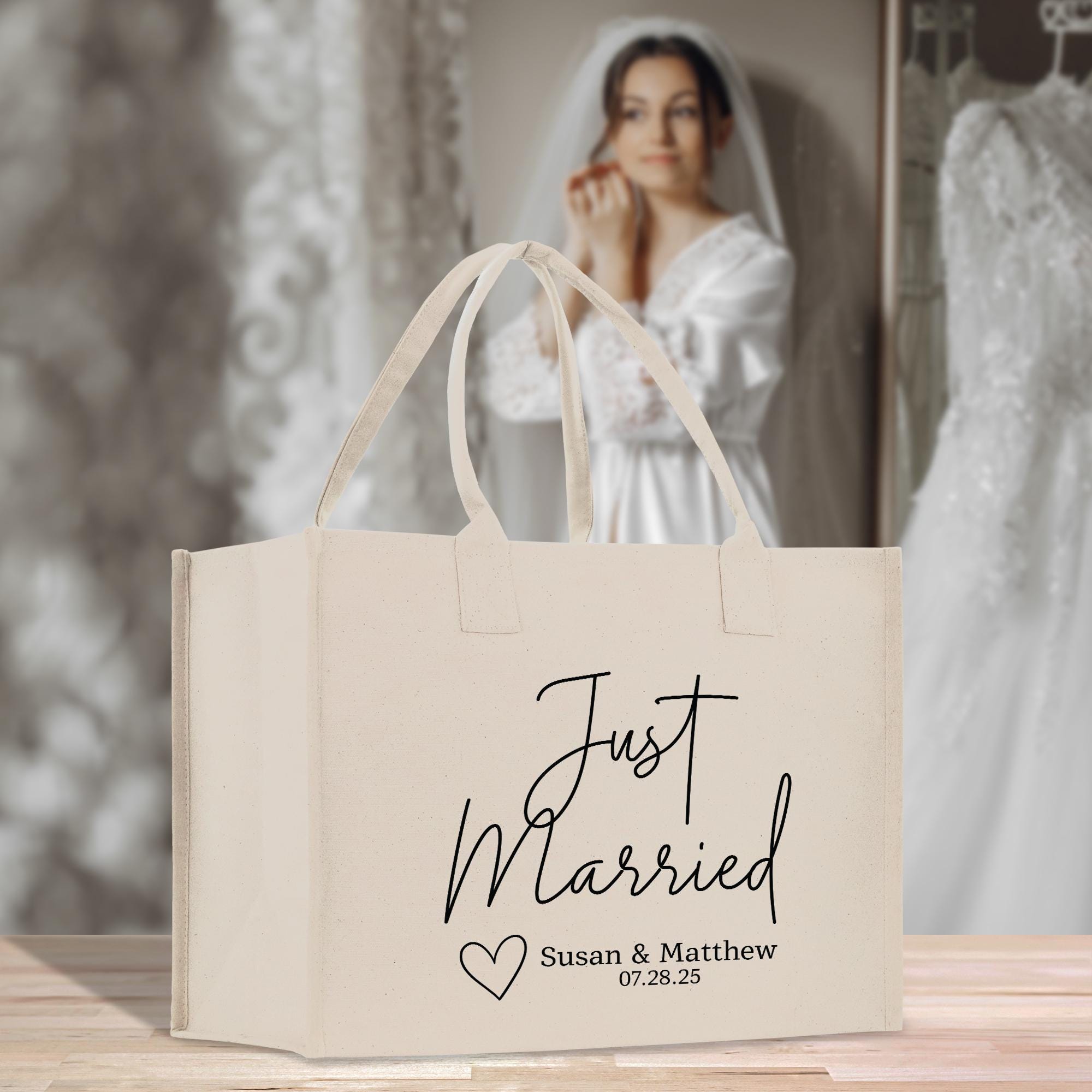 A beige tote bag with the text &quot;Just Married Susan & Matthew 07.28.25&quot; printed on it, placed on a wooden surface. In the background, a woman wearing a white wedding dress can be seen.