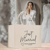 A beige tote bag with the text "Just Married Susan & Matthew 07.28.25" printed on it, placed on a wooden surface. In the background, a woman wearing a white wedding dress can be seen.