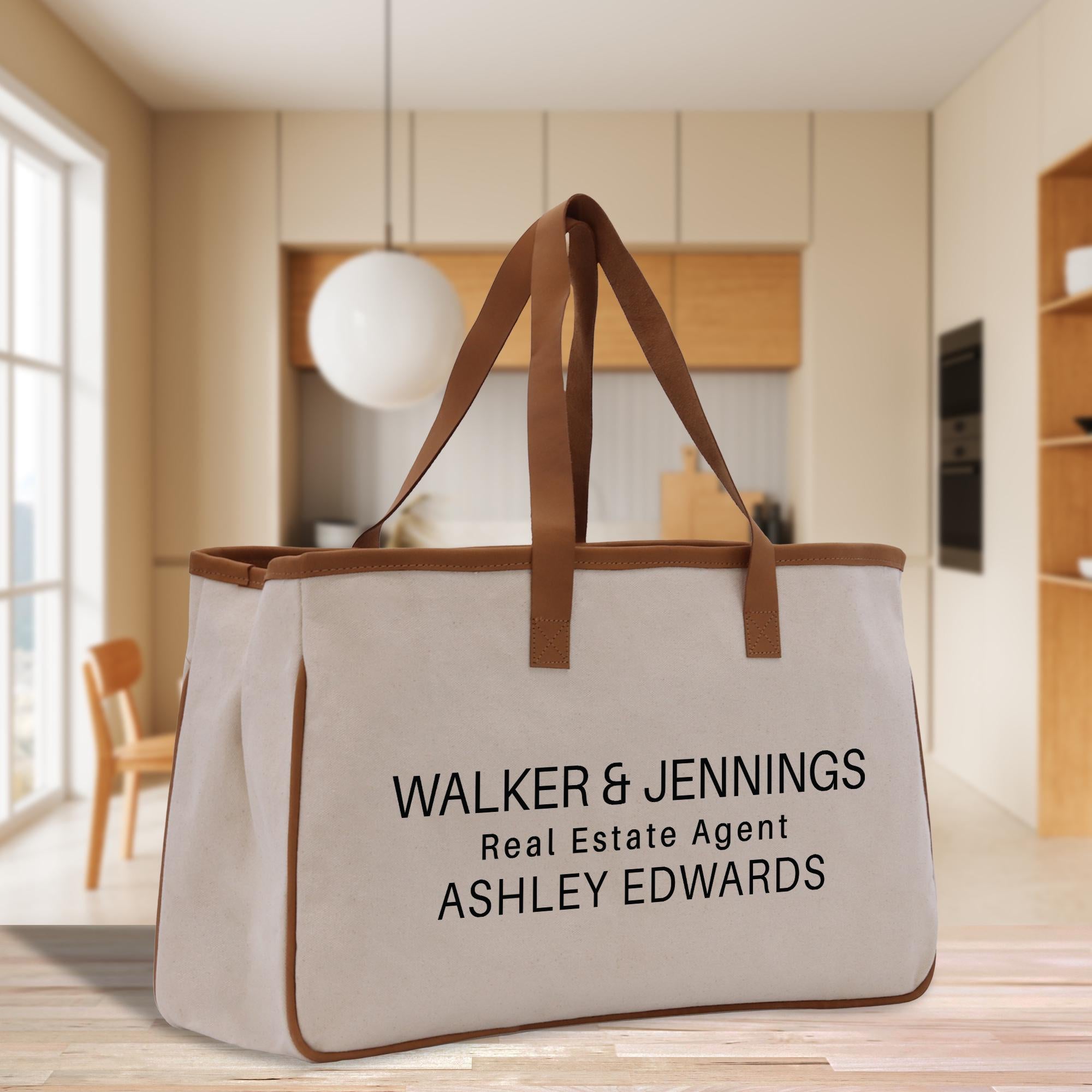 A beige canvas tote bag with the text &quot;WALKER & JENNINGS Real Estate Agent ASHLEY EDWARDS&quot; printed on it, placed on a wooden floor in a modern-looking room with a window and wooden furniture.