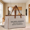 A beige canvas tote bag with the text "WALKER & JENNINGS Real Estate Agent ASHLEY EDWARDS" printed on it, placed on a wooden floor in a modern-looking room with a window and wooden furniture.