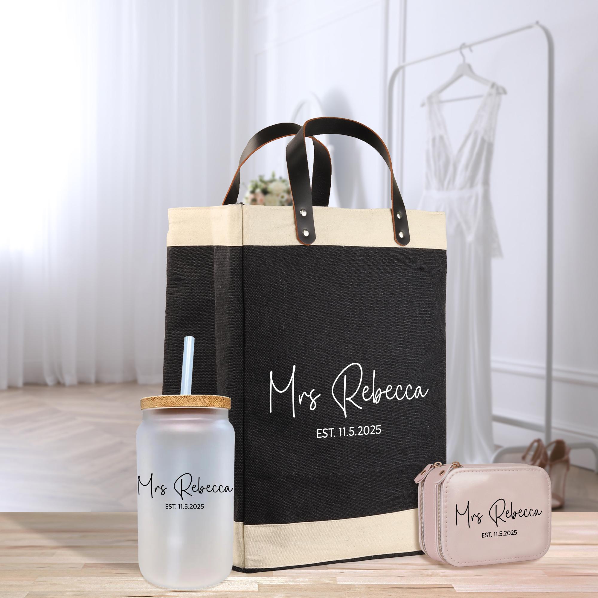 A black and beige tote bag with the text &quot;Mrs Rebecca EST. 11. 5. 2025&quot; printed on it, along with a matching black and beige cup with a straw and a small pink bag.