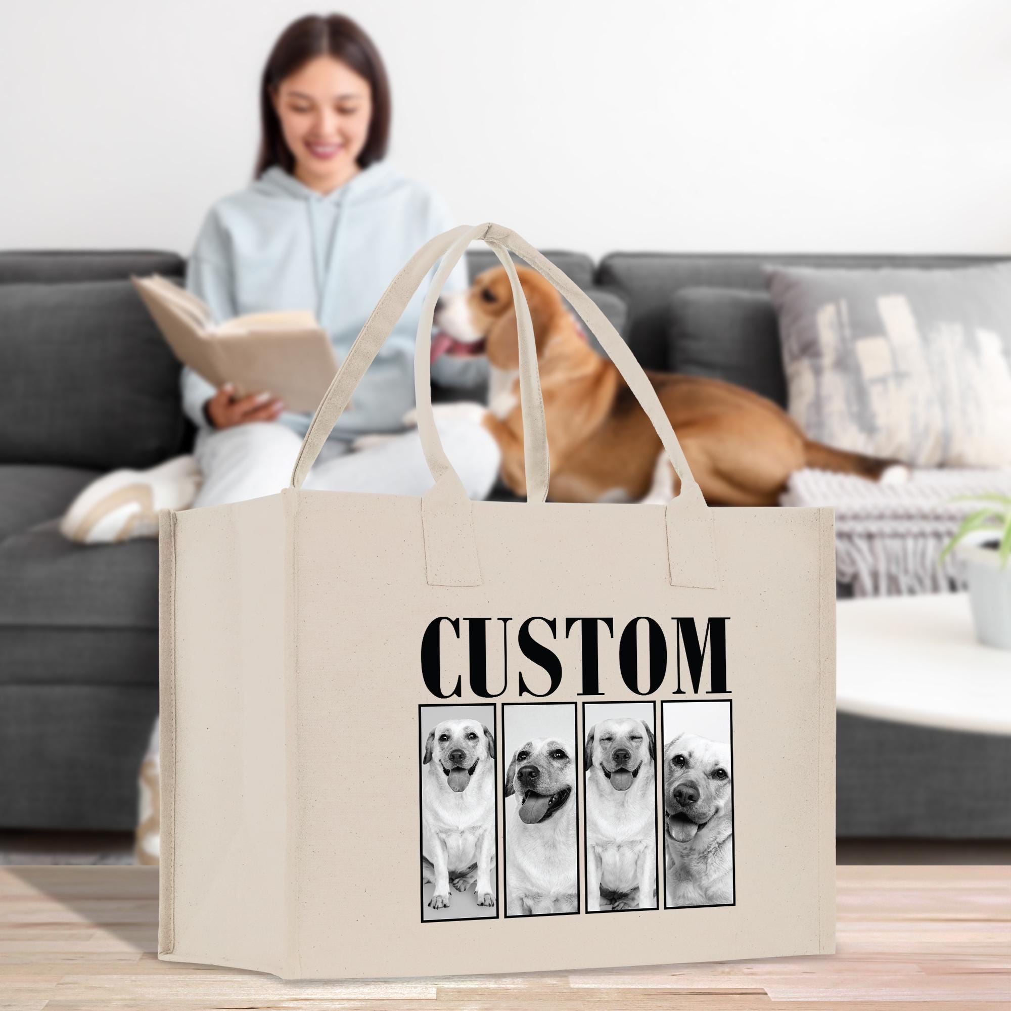 A woman is sitting on a couch, reading a book, while a dog is sitting next to her. In the foreground, there is a large canvas bag with a custom design featuring a collage of four dog portraits.