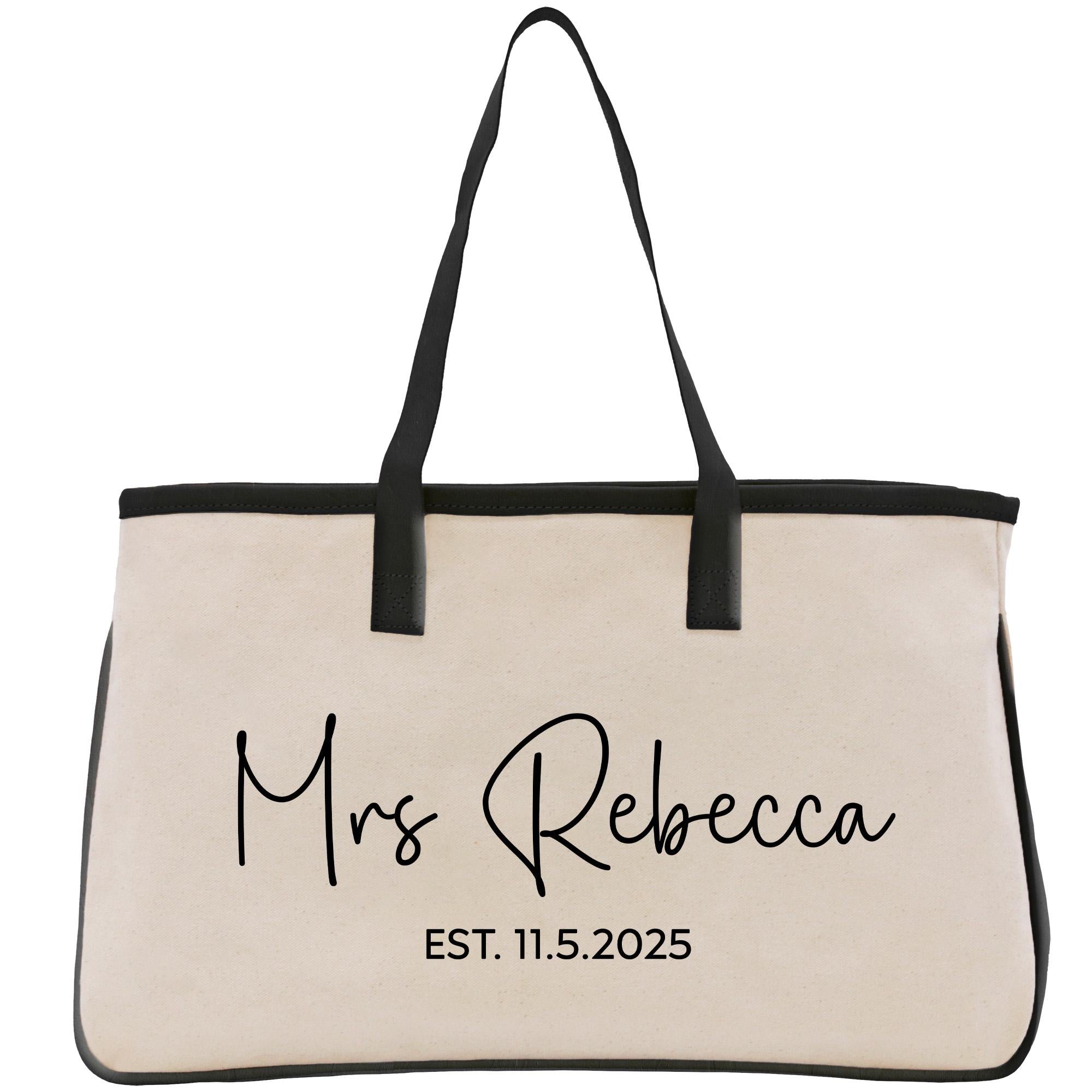 A beige canvas tote bag with black straps and handles, featuring personalized text and a date printed on it.
