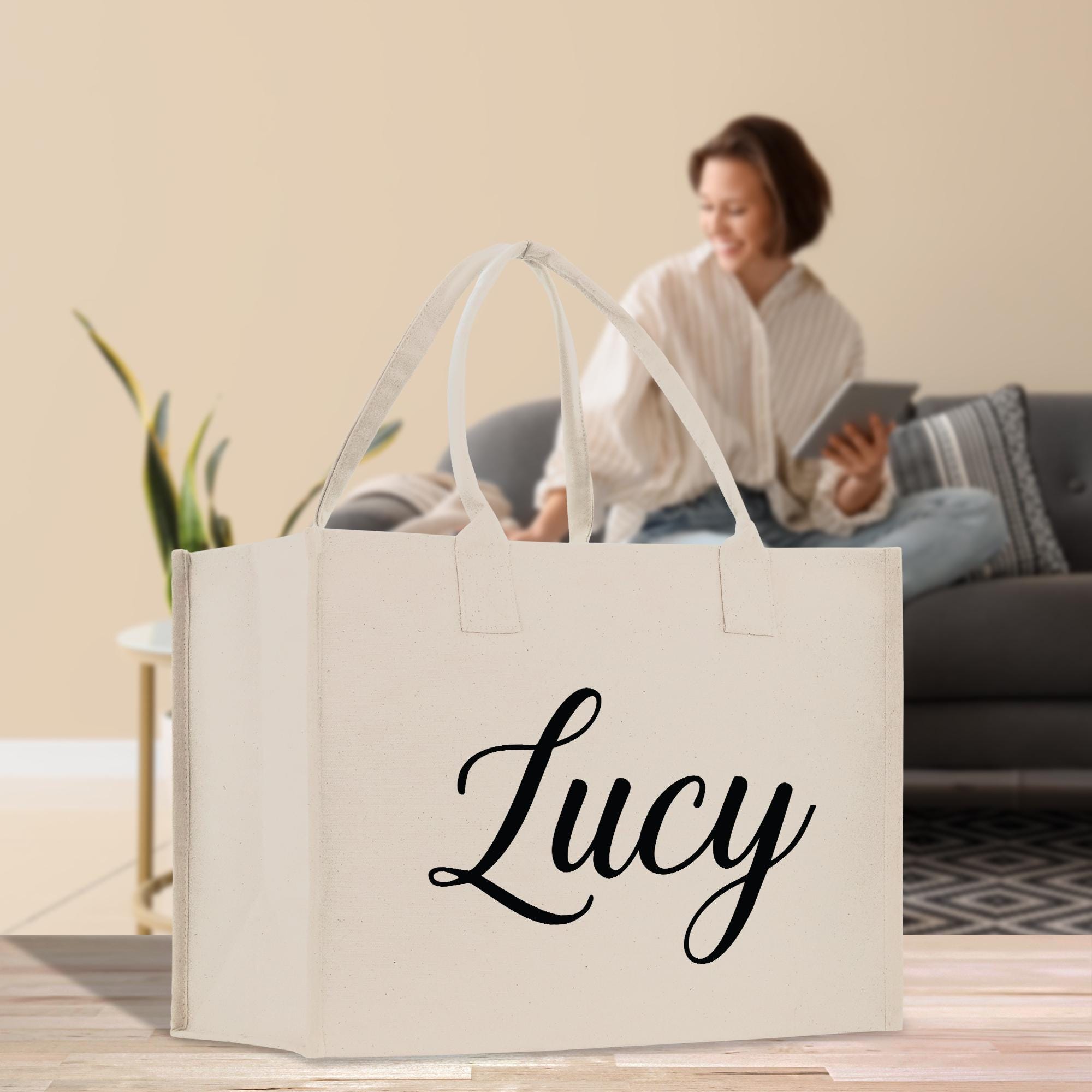 A white tote bag with the name &quot;Lucy&quot; printed on it, placed on a wooden floor in front of a woman sitting on a couch.