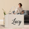 A white tote bag with the name "Lucy" printed on it, placed on a wooden floor in front of a woman sitting on a couch.