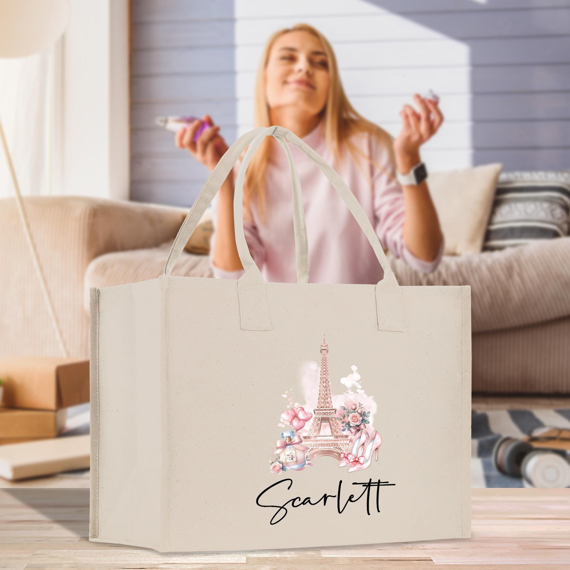 A woman is sitting on a couch in a living room, holding a phone and smiling. In front of her is a beige tote bag with a pink and white Eiffel Tower design and the name &quot;Scarflett&quot; printed on it.