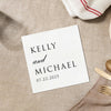 A white square card with black text is placed on a beige surface, along with a white napkin and a gold fork.