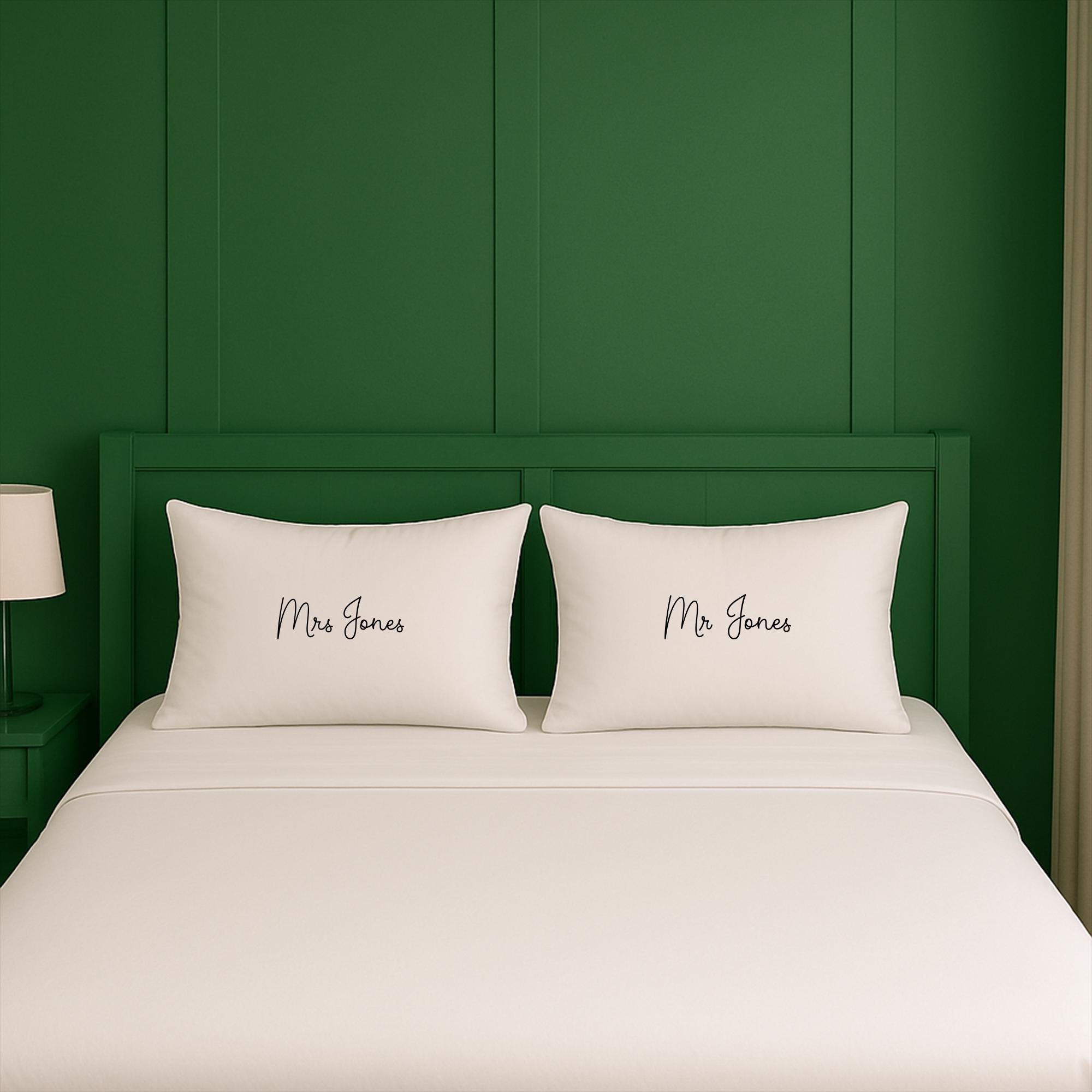 A white bed with two pillows, each with the name &quot;Mrs Jones&quot; written on it, against a green wall.