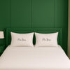A white bed with two pillows, each with the name "Mrs Jones" written on it, against a green wall.