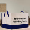 A white and blue tote bag with the text "Your custom wording here" printed on it, placed on a wooden surface.