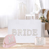 A white bag with the word "BRIDE" written on it, along with two smaller bags with the letter "M" on them, placed on a wooden surface.