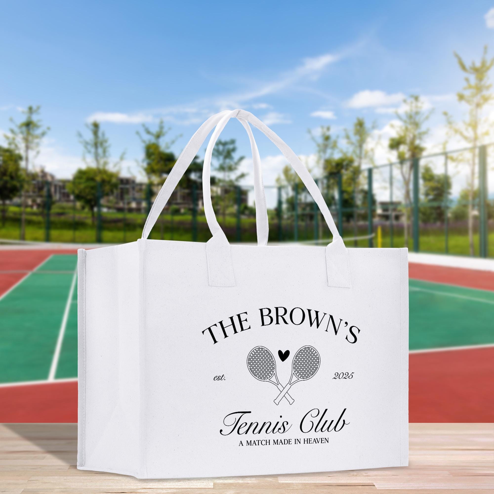 A white tote bag with text and graphics printed on it, placed on a wooden surface against a tennis court background.