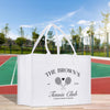 A white tote bag with text and graphics printed on it, placed on a wooden surface against a tennis court background.