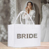 A white tote bag with the word "BRIDE" printed on it, placed on a wooden surface. In the background, there is a woman wearing a white wedding dress, visible through a mirror.