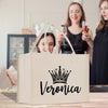 a white tote bag with the name "Veronica" printed on it, along with a crown design. In the background, there are two women standing and holding wine glasses, suggesting a social gathering or celebration.